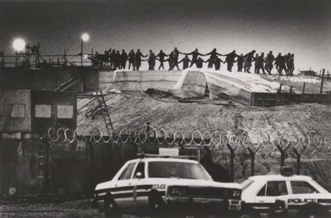 Black and white photograph. A few dozen women dance with hands joined in a ring on a mound. They are black silhouettes against a darkened sky. Two police cars are parked below.