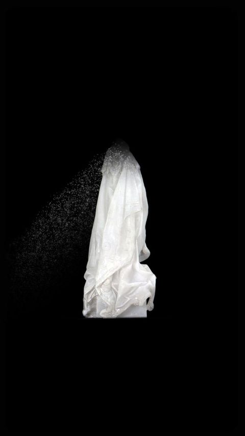 Video still. A white cloak hangs as if over someone’s shoulders against a black background. The top dissolves into white specs and floats down and to our left.