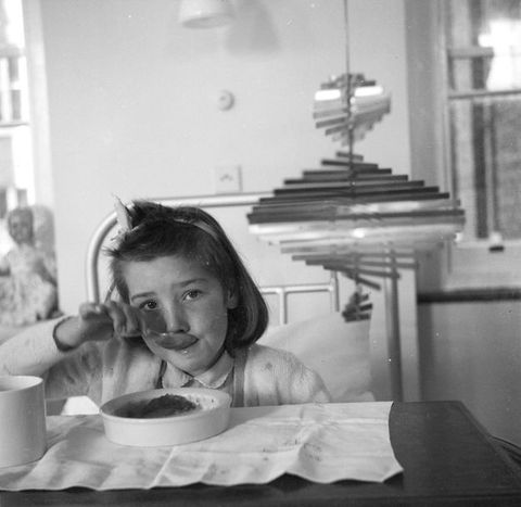 Black and white photograph. Child seated at a table takes a big bite from a spoon, looking at us with a curious expression. They are seated next to and below a rotating corkscrew-shaped metal mobile sculpture that's wide on the bottom and narrow at the top.