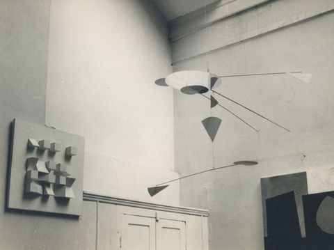 Exhibition. Abstract metal mobile sculpture with geometric shapes hangs from the ceiling next to a square-shaped panel fixed to the wall that contains a loose four by four grid of square and triangular blocks.