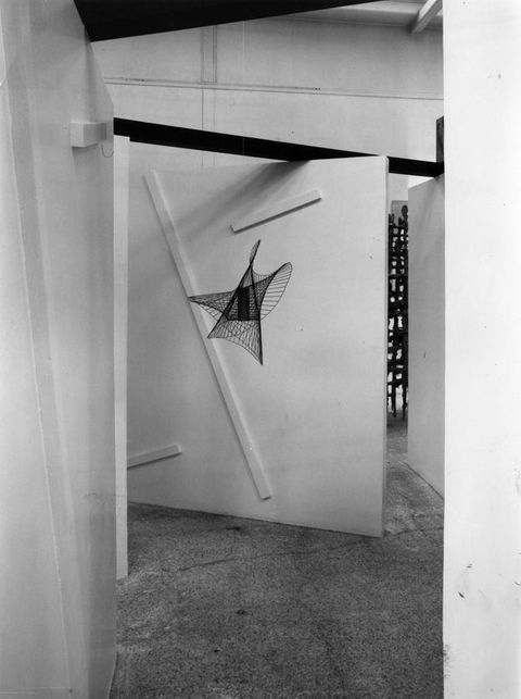 Exhibition. Black and white photograph of a view down an angled corridor towards a metal mobile sculpture that forms an abstract three-dimensional star shape with five curved points.