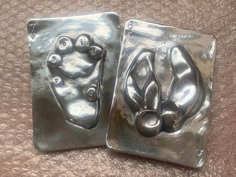 Metalwork. Two shiny silver plates with rounded corners like playing cards lie against bubble wrap. The seven of hearts to our left has a lion’s paw in relief. Letters on the toe pads together read 'spoils'. Next to it, the eight of clubs has two disembodied bull’s ears.