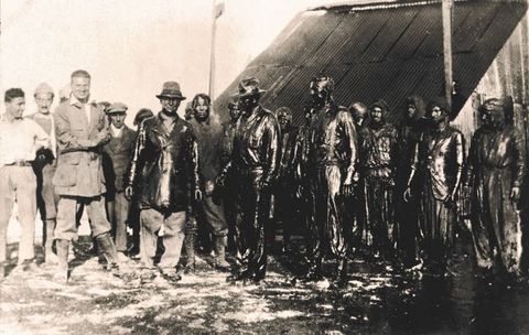 Black and white photograph. Nineteen men stand in front of a structure with s steeply pitched roof. Some of the six standing to our left smile. The rest are covered completely in slick black oil. A pool of oil surrounds their feet to our right.