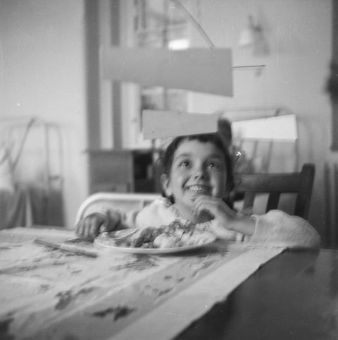 Black and white photograph. Child smiles happily up at a mobile sculpture from a dining table. The mobile consists of individual white trapezoidal shapes hanging from delicate strings attached to narrow metal supports above.