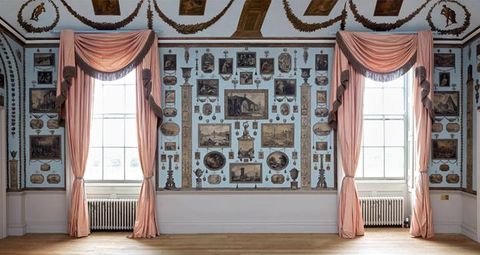 Installation. A pale blue wall has two windows draped with rose pink curtains. Prints are carefully and tidily arranged in the spaces between and around the windows.