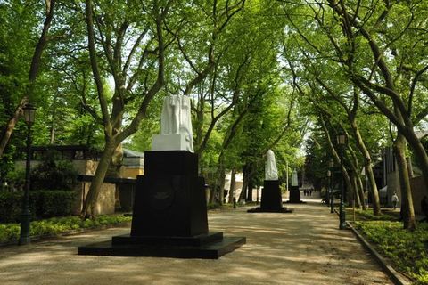 Public art. Three black plinths in a tree-covered lane hold what appear to be fragments of white statues. The statue closest to us seems broken off above the hips.